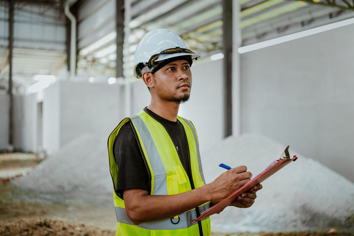 Supervisor in a helmet and safety vest writing on a paper form on a clipboard in a plant