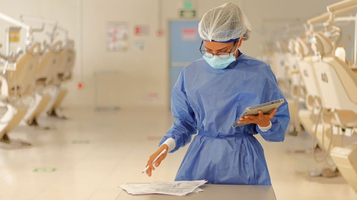 Medical staff in a mask and protective gown checking forms while holding a tablet